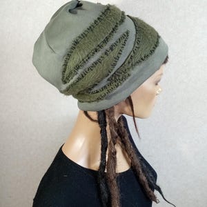 May include: An olive green beanie hat featuring a textured, fuzzy fabric overlay in a darker shade of green. The hat has a rounded crown and a wide, fitted band that sits snugly around the head, showcasing a layered, multi-dimensional appearance.