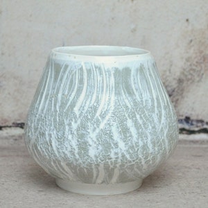 Peut inclure: Un vase en céramique blanche avec un glaçage gris clair et une finition craquelée. Le vase a un corps arrondi et une large ouverture.