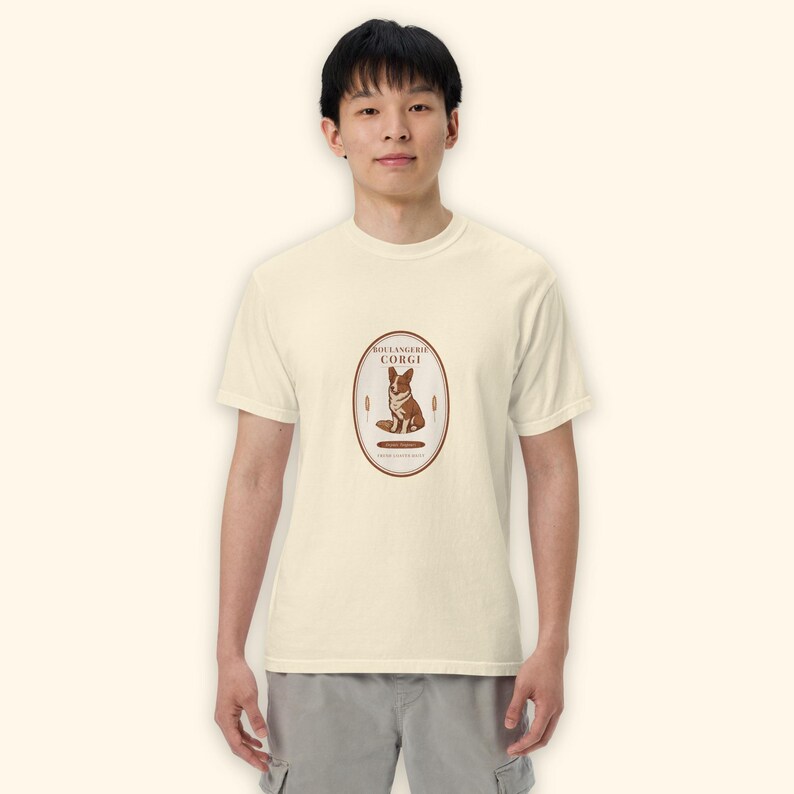 May include: A cream-colored short-sleeve t-shirt featuring a central oval graphic. The design includes a brown and white corgi illustration, the text "BOULANGERIE CORGI," and wheat stalks. The shirt has a crew neckline and a standard fit.