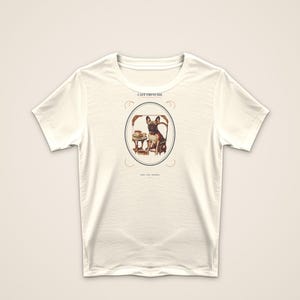 Puede incluir: Una camiseta de manga corta de color crema con un gr&aacute;fico central de un bulldog franc&eacute;s sentado en una mesa peque&ntilde;a con una taza y un plato. El texto CAF&Eacute; FRENCHIE est&aacute; impreso sobre el marco ovalado y hay texto m&aacute;s peque&ntilde;o debajo.