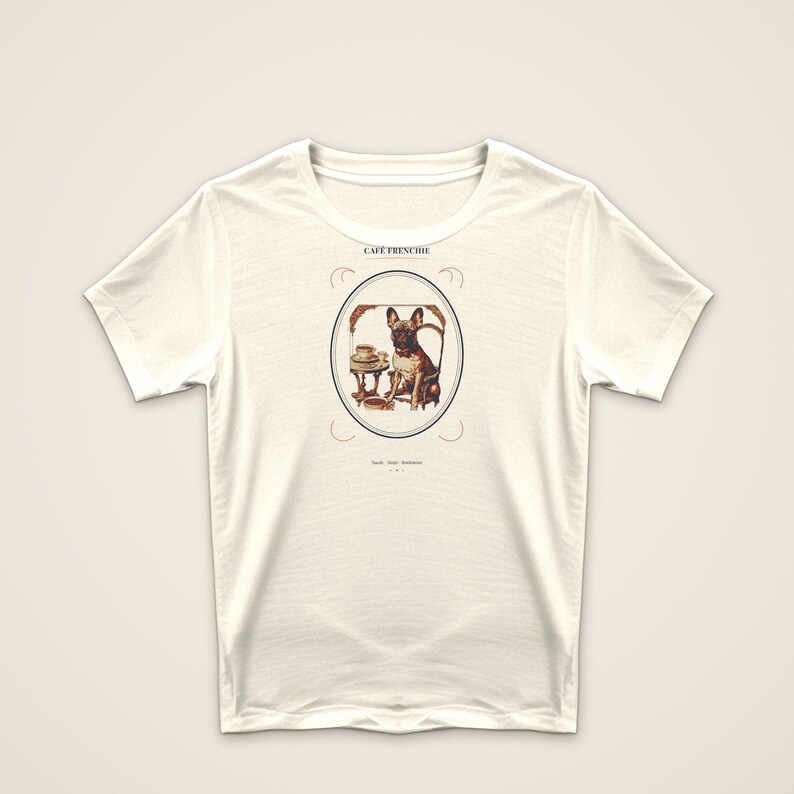 Puede incluir: Una camiseta de manga corta de color crema con un gr&aacute;fico central de un bulldog franc&eacute;s sentado en una mesa peque&ntilde;a con una taza y un plato. El texto CAF&Eacute; FRENCHIE est&aacute; impreso sobre el marco ovalado y hay texto m&aacute;s peque&ntilde;o debajo.