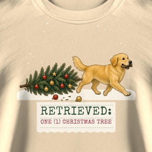 May include: A cream-colored t-shirt featuring a golden retriever carrying a decorated Christmas tree in its mouth. The graphic includes the text "RETRIEVED: ONE (1) CHRISTMAS TREE" in green and red lettering, set against a snowy background.