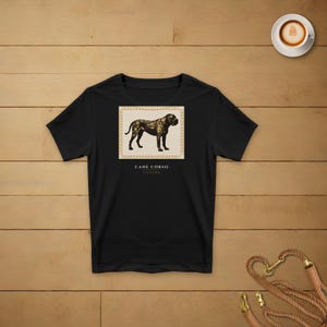 May include: A black short-sleeved t-shirt featuring a rectangular graphic on the chest. The graphic displays a brown and black Cane Corso dog illustration inside a bordered frame, with the text CANE CORSO CUSTOS printed directly below the image.