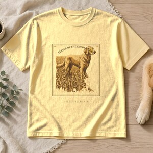 May include: A pale yellow short-sleeve t-shirt featuring a rectangular graphic print of a golden retriever standing in tall grass. The text above the dog reads KEEPER OF THE GOLDEN HOUR and the text below the dog reads GOLDEN RETRIEVER.