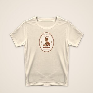 May include: A cream-colored short-sleeve t-shirt featuring a central oval graphic. The design includes a brown and white Corgi illustration, the text "BOULANGERIE CORGI," and wheat stalks. The print is rendered in shades of brown on the fabric.