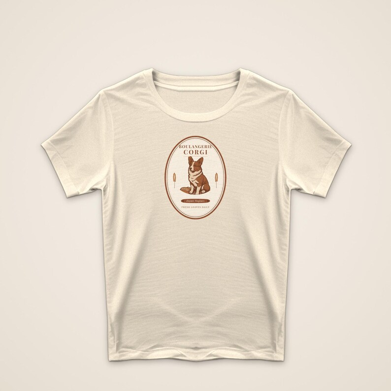 May include: A cream-colored short-sleeve t-shirt featuring a central oval graphic. The design includes a brown and white Corgi illustration, the text "BOULANGERIE CORGI," and wheat stalks. The print is rendered in shades of brown on the fabric.