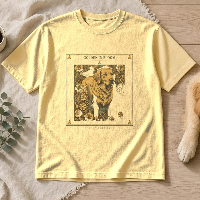 May include: A pale yellow short-sleeve t-shirt featuring a rectangular graphic print of a Golden Retriever surrounded by roses. The text "GOLDEN IN BLOOM" appears above the dog, and "GOLDEN RETRIEVER" is printed below the illustration.