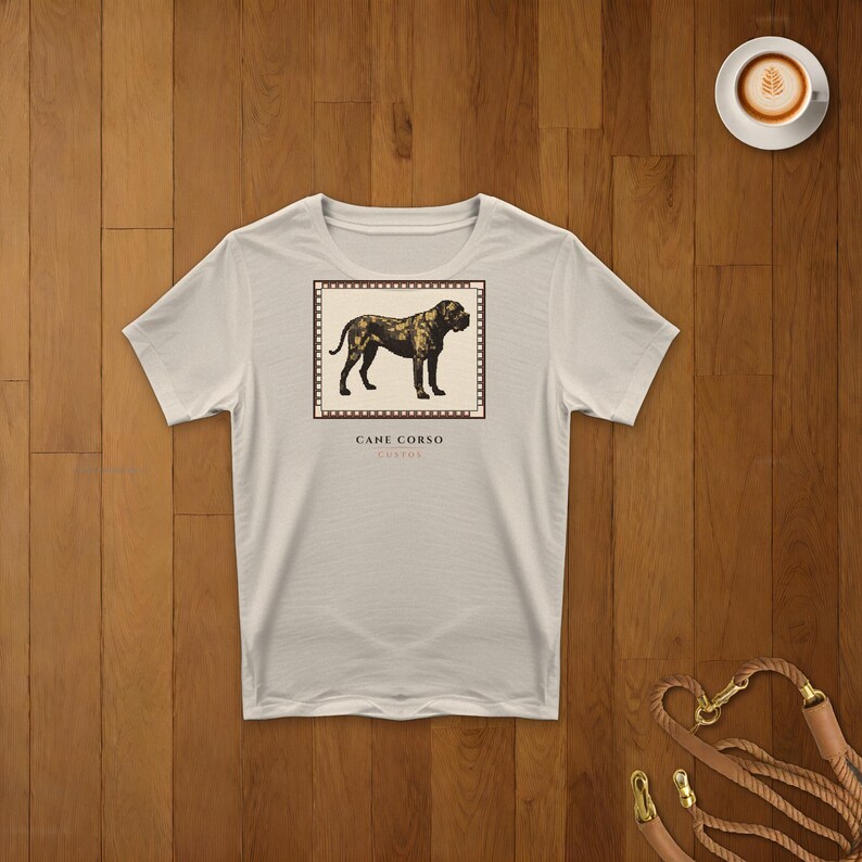 May include: A light beige short-sleeved t-shirt featuring a rectangular graphic of a dark brown Cane Corso dog standing in profile. The graphic is framed by a border resembling film strip perforations, with the text CANE CORSO printed below.