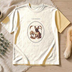 Puede incluir: Una camiseta de manga corta color crema con mangas amarillas. El centro presenta un gr&aacute;fico de un bulldog franc&eacute;s sentado en una mesa con una taza de caf&eacute;, encerrado en un &oacute;valo con el texto CAF&Eacute; FRENCHIE impreso sobre la ilustraci&oacute;n.
