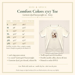 Puede incluir: Una camiseta color marfil Comfort Colors 1717 de tejido pesado con un gr&aacute;fico de un perro en un marco ovalado. La imagen incluye una tabla de tallas con longitudes y anchos de pecho en pulgadas, detalles de algod&oacute;n e instrucciones de cuidado.