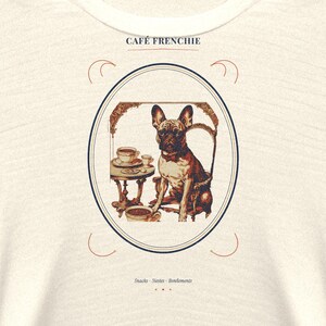 Puede incluir: Una camiseta de color crema con un gr&aacute;fico de un bulldog franc&eacute;s sentado en una silla junto a una mesa con caf&eacute; y aperitivos. El texto CAF&Eacute; FRENCHIE aparece sobre el marco ovalado y las palabras Snacks &middot; Siestes &middot; Ronflements debajo.