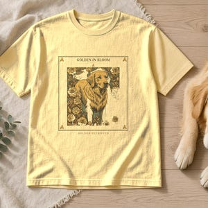 May include: A pale yellow t-shirt featuring a rectangular graphic print of a Golden Retriever dog surrounded by floral illustrations. The text "GOLDEN IN BLOOM" appears above the dog, and the words "GOLDEN RETRIEVER" are printed below the graphic.