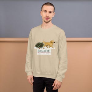 May include: A beige crewneck sweatshirt featuring a graphic of a golden retriever dog carrying a decorated Christmas tree in its mouth. Below the dog, the text reads: RETRIEVED: ONE (1) CHRISTMAS TREE. The design includes small scattered ornaments.