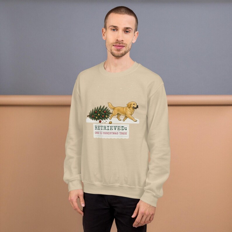 May include: A beige crewneck sweatshirt featuring a graphic of a golden retriever dog carrying a decorated Christmas tree in its mouth. Below the dog, the text reads: RETRIEVED: ONE (1) CHRISTMAS TREE. The design includes small scattered ornaments.