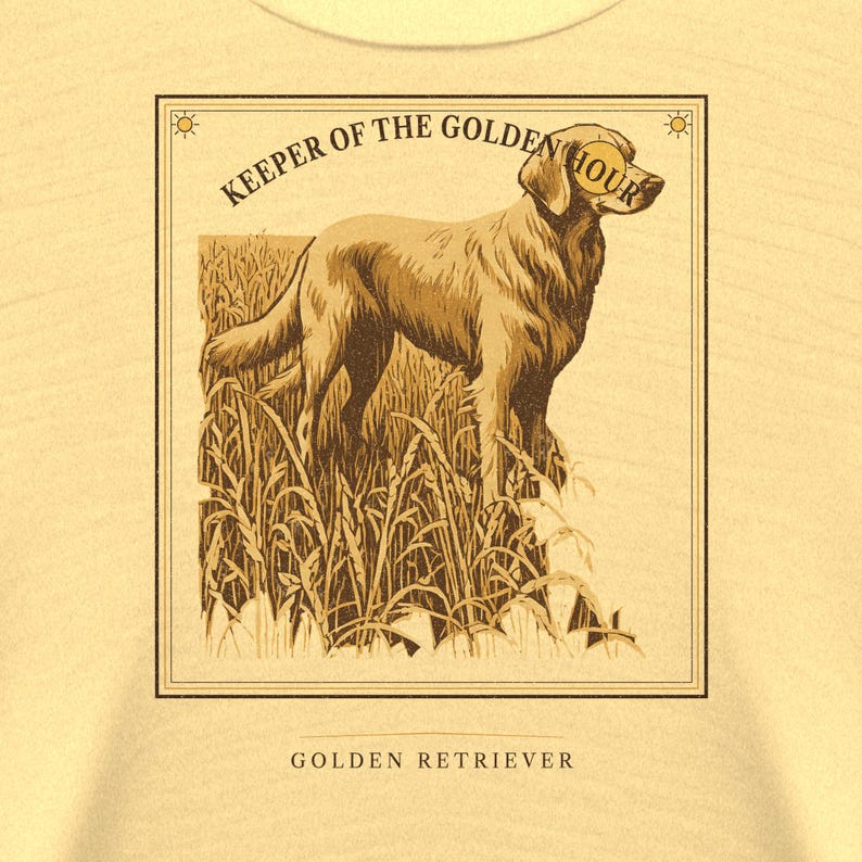 May include: A light yellow t-shirt featuring a rectangular graphic of a golden retriever standing in a field of tall grass. The text KEEPER OF THE GOLDEN HOUR arches above the dog, and the words GOLDEN RETRIEVER are printed below the frame.