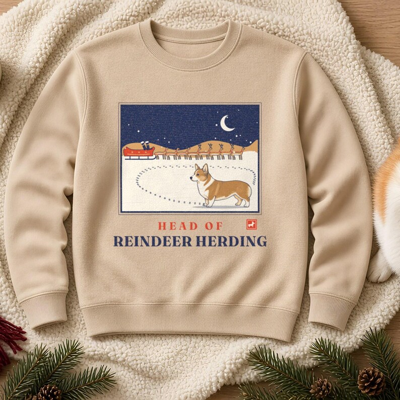 May include: A beige crewneck sweatshirt featuring a graphic of a Corgi standing in snow with a dotted path behind it. Above the dog is a night scene with a red sleigh pulled by reindeer. The text reads HEAD OF REINDEER HERDING in blue letters.