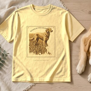 May include: A pale yellow short-sleeve t-shirt featuring a dark brown rectangular graphic print of a dog standing in tall grass. The text above the dog reads KEEPER OF THE GOLDEN HOUR and the text below the dog reads GOLDEN RETRIEVER.