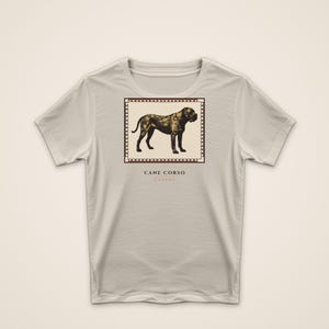 May include: A light beige short-sleeve t-shirt featuring a square graphic of a dark brown Cane Corso dog inside a film strip border. Below the graphic, the text reads CANE CORSO and CUSTOS in a dark brown font centered on the chest area.