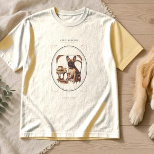 Puede incluir: Una camiseta de manga corta de color crema con un gr&aacute;fico central de un bulldog franc&eacute;s sentado a una mesa con una taza de caf&eacute;. El texto "CAFE FRENCHIE" est&aacute; impreso sobre el perro en una fuente fina y oscura dentro de un marco ovalado.