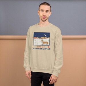 May include: A beige crewneck sweatshirt featuring a graphic illustration of a Corgi standing in snow with a sleigh and reindeer in the background under a crescent moon. The text reads HEAD OF REINDEER HERDING in blue capital letters.