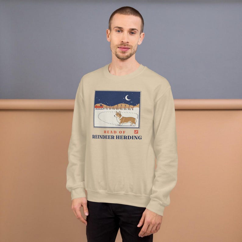 May include: A beige crewneck sweatshirt featuring a graphic illustration of a Corgi standing in snow with a sleigh and reindeer in the background under a crescent moon. The text reads HEAD OF REINDEER HERDING in blue capital letters.