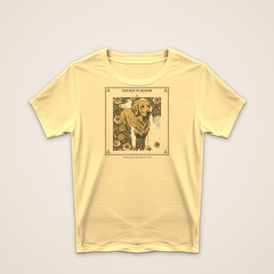 May include: A pale yellow short-sleeve t-shirt featuring a rectangular graphic print of a golden retriever surrounded by roses. The text "GOLDEN IN BLOOM" appears above the dog, and "GOLDEN RETRIEVER" is printed below the illustration.