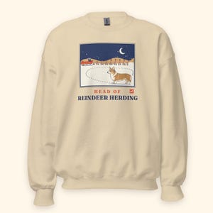 May include: A cream-colored crewneck sweatshirt featuring a graphic illustration of a Corgi standing in snow with a red sleigh and reindeer in the background under a crescent moon. The text reads HEAD OF REINDEER HERDING in blue and red.