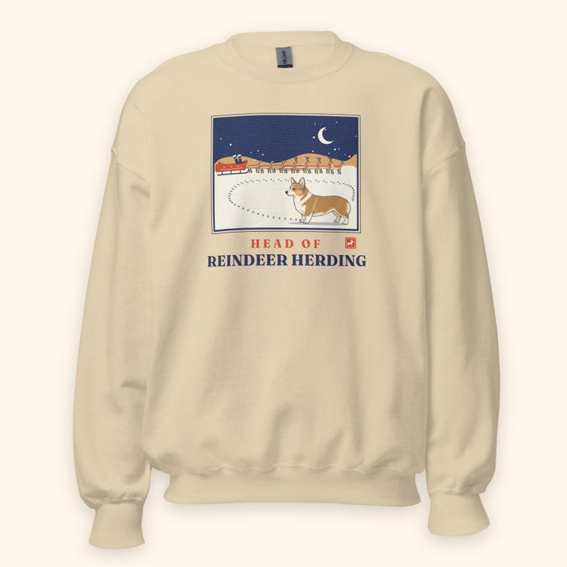 May include: A cream-colored crewneck sweatshirt featuring a graphic illustration of a Corgi standing in snow with a red sleigh and reindeer in the background under a crescent moon. The text reads HEAD OF REINDEER HERDING in blue and red.