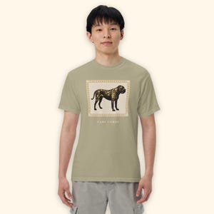 May include: A sage green short-sleeve t-shirt featuring a rectangular graphic of a Cane Corso dog in shades of brown and black. The graphic is framed by a light-colored border and includes the text CANE CORSO printed in capital letters below it.