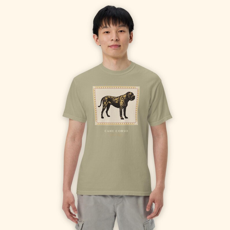 May include: A sage green short-sleeve t-shirt featuring a rectangular graphic of a Cane Corso dog in shades of brown and black. The graphic is framed by a light-colored border and includes the text CANE CORSO printed in capital letters below it.