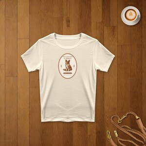 May include: A cream-colored short-sleeve t-shirt featuring a central oval graphic. The graphic contains a brown illustration of a Corgi dog, the text "BOULANGERIE CORGI" in a dark brown font, and small wheat stalks on either side of the dog.