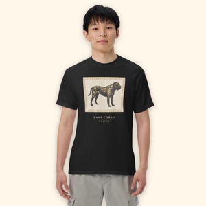 May include: A black short-sleeve t-shirt featuring a rectangular graphic of a Cane Corso dog in shades of brown and gold. The graphic is framed by a light-colored border and includes the text CANE CORSO CUSTOS printed in black below the image.