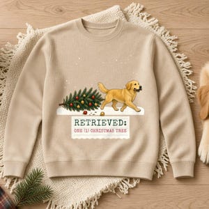 May include: A beige crewneck sweatshirt featuring a graphic of a golden retriever dog carrying a decorated Christmas tree. Below the dog, the text reads: RETRIEVED: ONE (1) CHRISTMAS TREE. The design includes small white dots representing falling snow.