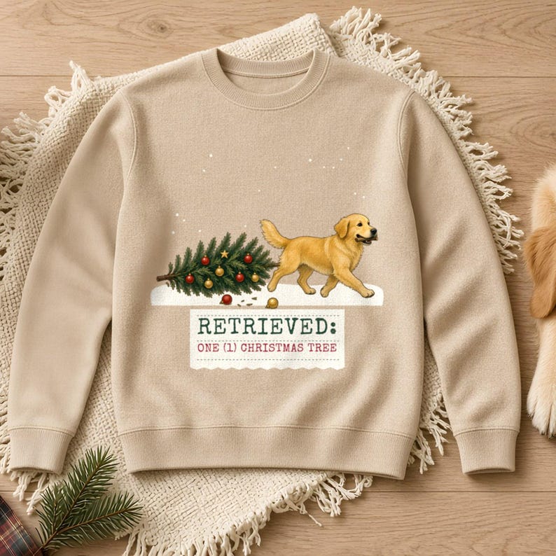 May include: A beige crewneck sweatshirt featuring a graphic of a golden retriever dog carrying a decorated Christmas tree. Below the dog, the text reads: RETRIEVED: ONE (1) CHRISTMAS TREE. The design includes small white dots representing falling snow.