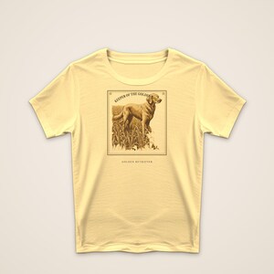 May include: A pale yellow short-sleeve t-shirt featuring a rectangular graphic print of a Golden Retriever standing in tall grass. The text above the dog reads KEEPER OF THE GOLDEN HOUR and the text below the graphic reads GOLDEN RETRIEVER.