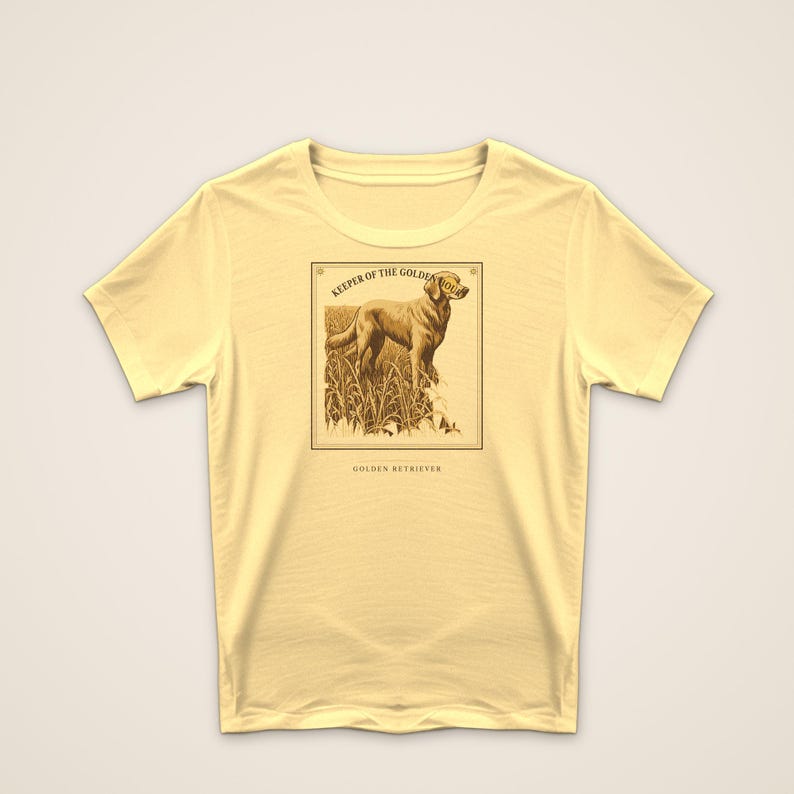May include: A pale yellow short-sleeve t-shirt featuring a rectangular graphic print of a Golden Retriever standing in tall grass. The text above the dog reads KEEPER OF THE GOLDEN HOUR and the text below the graphic reads GOLDEN RETRIEVER.