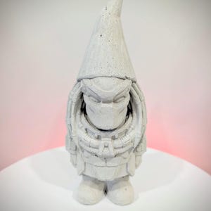 May include: A light gray concrete garden gnome figurine featuring a textured surface and a pointed hat. The gnome is sculpted with detailed armor plating across the chest and head, resembling a science fiction character design.