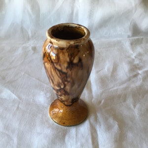 May include: A small, brown ceramic vase with a flared top and a pedestal base. The vase has a glossy finish with a marbled pattern of dark brown and tan colors. The base is a solid, rounded shape.