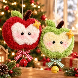 May include: Two plush apple ornaments hanging by strings. One is red with a white center, the other is green with a light yellow center. Both feature embroidered eyes, pink cheeks, a small yellow star, a checkered bow, and a metal bell.