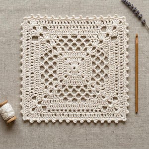 May include: A square off-white crochet doily featuring a central starburst pattern, intricate openwork mesh, and a scalloped border. A wooden crochet hook and a small spool of matching off-white yarn are positioned beside the square textile piece.