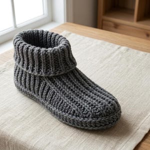 Puede incluir: Una bota de estar por casa tejida en color gris oscuro que presenta una textura acanalada gruesa en todo el cuerpo y un puño ancho doblado en el tobillo. El calzado está confeccionado con un patrón de punto grueso, con forma estructurada.