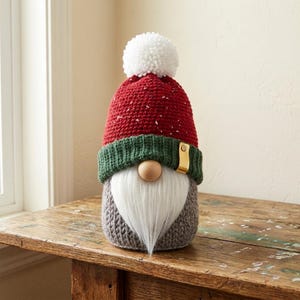 May include: A gnome figurine featuring a gray knitted body, a long white faux fur beard, and a round wooden nose. The gnome wears a red crocheted hat with white speckles, a green ribbed brim, a white pom-pom, and a small gold-colored metal tag.