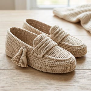 May include: A pair of beige crochet loafers featuring a textured knit construction, a rounded toe, and a decorative tassel detail attached to the side of the shoe. The slip-on footwear has a structured silhouette with a visible stitch pattern.