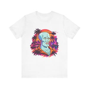 May include: A white t-shirt featuring a graphic print of a blue, humanoid character with a prominent jawline, wearing an orange shirt. The background displays a sunset, palm trees, a helicopter, a sports car, and a city skyline.