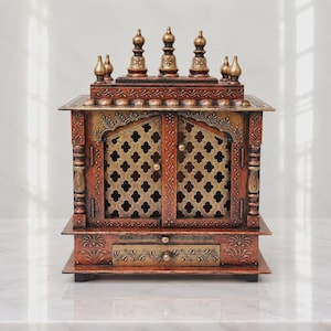 May include: A wooden home temple cabinet featuring a reddish-brown finish with gold-toned accents. The structure includes two doors with intricate lattice cutouts, a small pull-out drawer at the base, and five decorative spires on top.