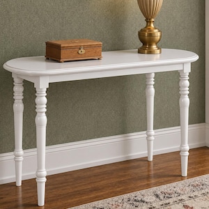 May include: A white console table with a smooth, oval-shaped top and four turned, spindle-style legs. A rectangular wooden box with a gold-toned metal latch and a gold-toned table lamp are placed on the surface of the white console table.