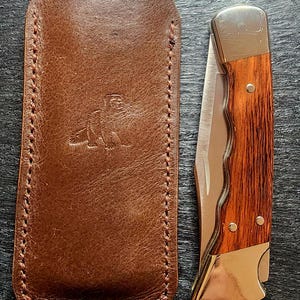 May include: A folding pocket knife with a wooden handle and silver metal bolsters, positioned next to a brown leather sheath featuring a stamped animal emblem and stitched edges. The knife has a curved, metallic blade edge visible.