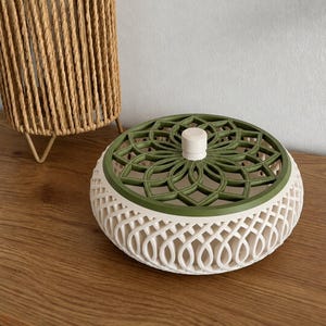 Può includere: Un contenitore rotondo con coperchio, caratterizzato da una base bianca con motivo a reticolo e un coperchio verde oliva scuro con un design geometrico traforato a forma di fiore. Un piccolo pomello cilindrico bianco è centrato sul coperchio.
