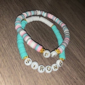 May include: Two beaded bracelets with the words "Strong" and "Fly" spelled out in gold letters on white beads. The bracelets are made with turquoise, pink, white, and gray beads.