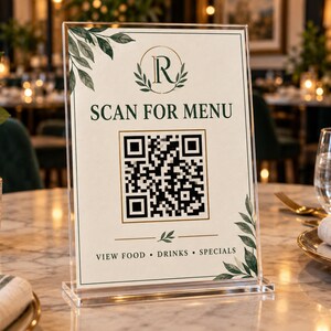 May include: A clear acrylic sign holder containing a rectangular menu card with a white background, green leaf illustrations, a central QR code, and the text SCAN FOR MENU, VIEW FOOD, DRINKS, SPECIALS, and a circular logo with the letter R.